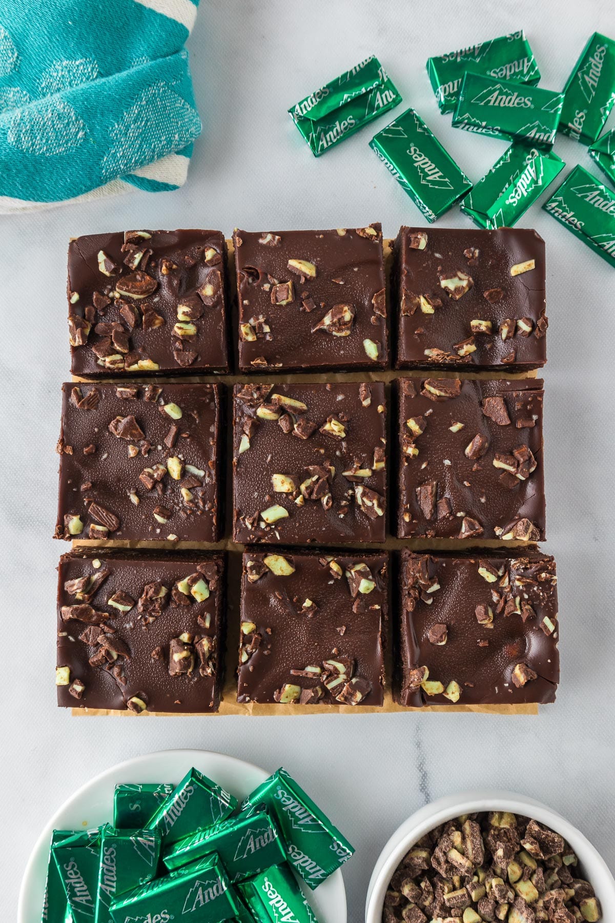 Nine square pieces of chocolate Andes mint brownies topped with crumbled mint candies on a cutting board with more foil wrapped Andes mints nearby on the counter.