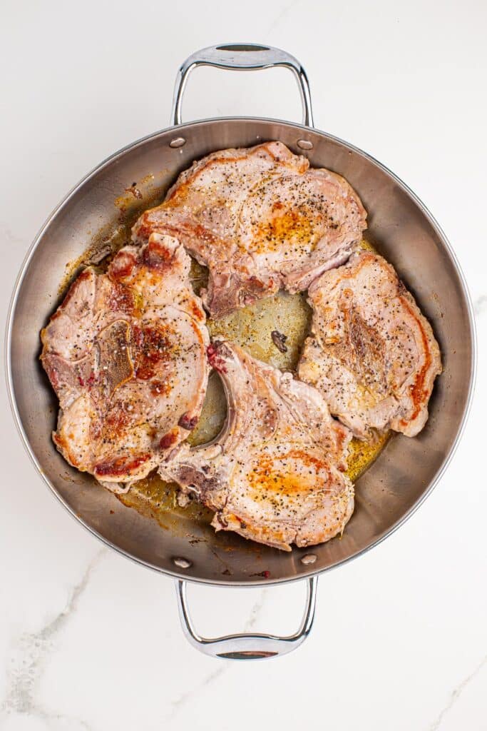 Four seasoned pork chops seasoned and seared to a golden brown with cooking liquids in the bottom of a skillet.