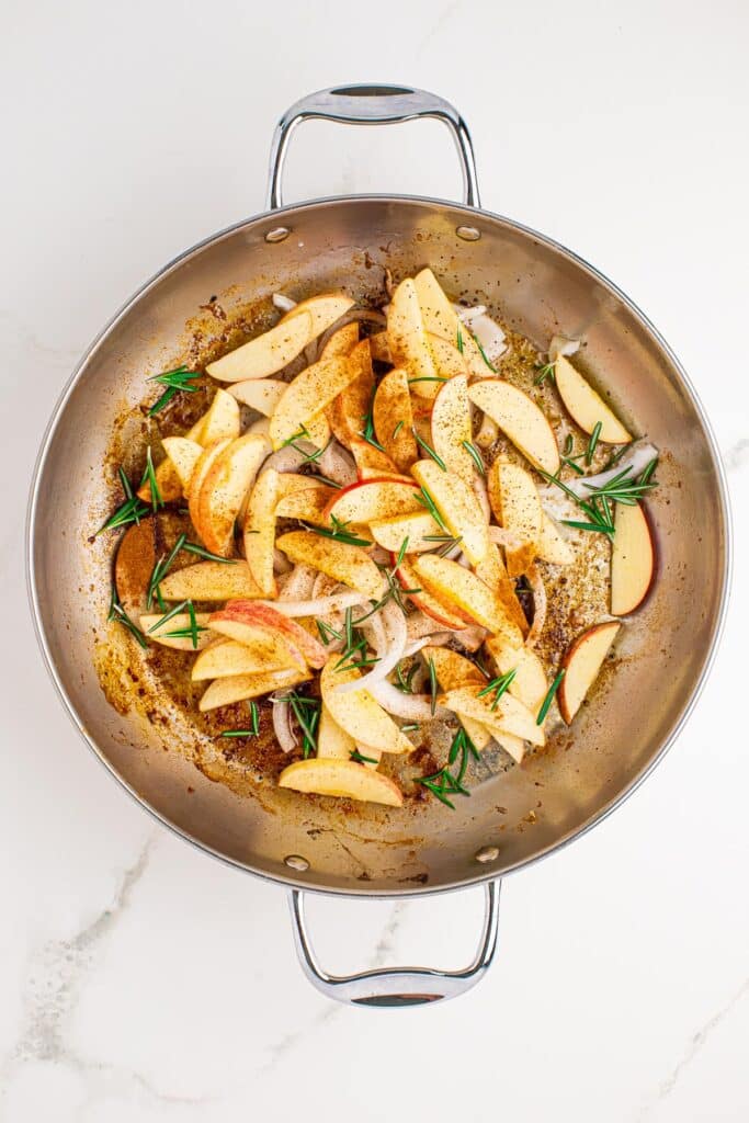 A skillet full of raw apples, onions, rosemary and seasoning.