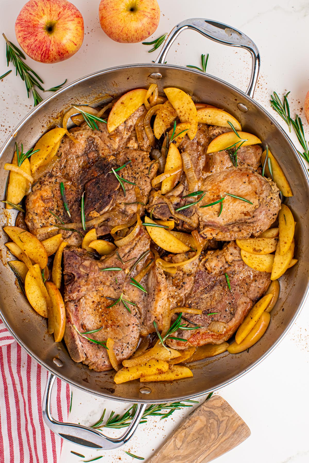 A stainless steel skillet full of cooked pork chops and apples topped with fresh rosemary.