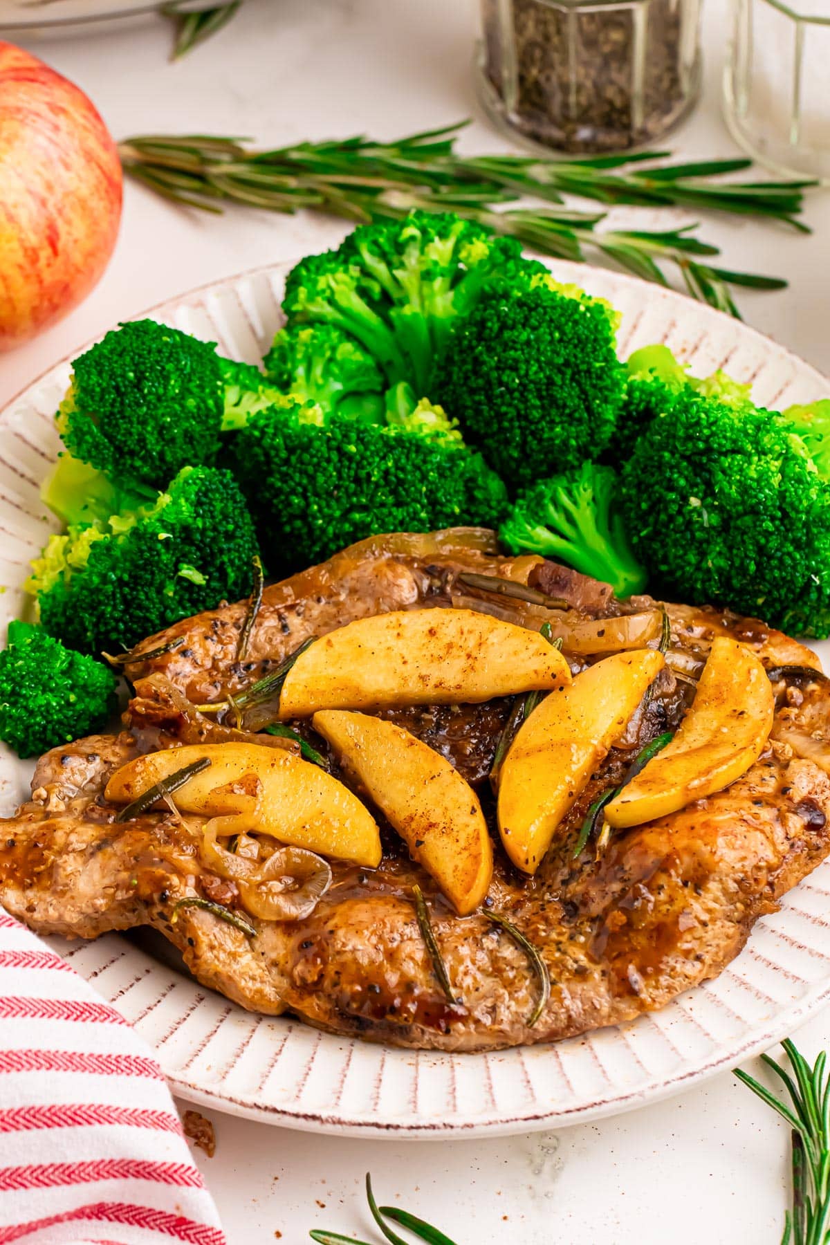 A plate of cooked pork chops topped with cooked apple slices and an apple butter sauce served with steamed broccoli on a table.