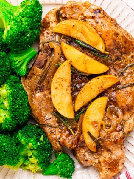 A plate with a cooked apple butter pork chop topped with cooked apple slices next to steamed broccoli on a plate.