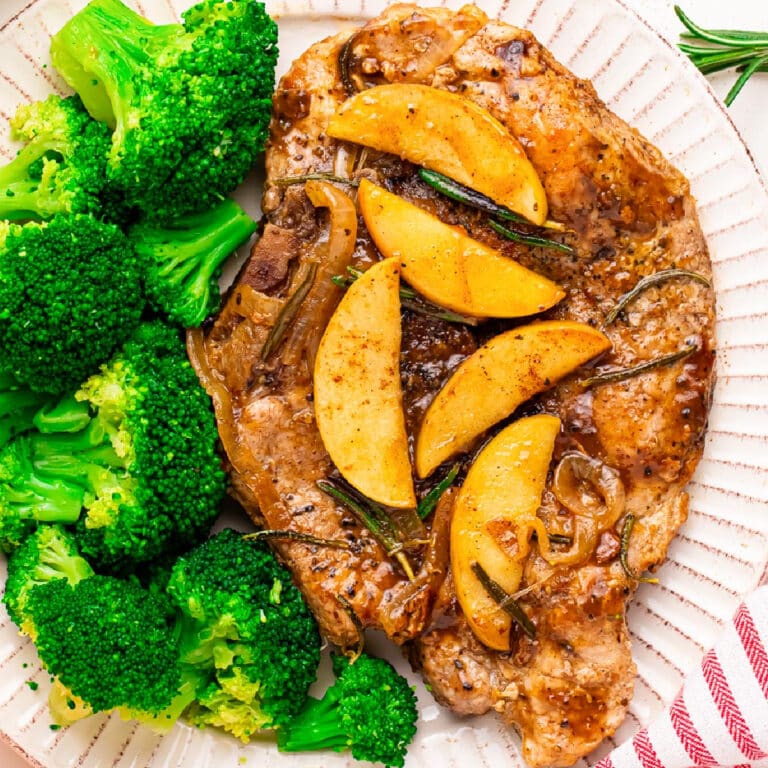 A plate with a cooked apple butter pork chop topped with cooked apple slices next to steamed broccoli on a plate.