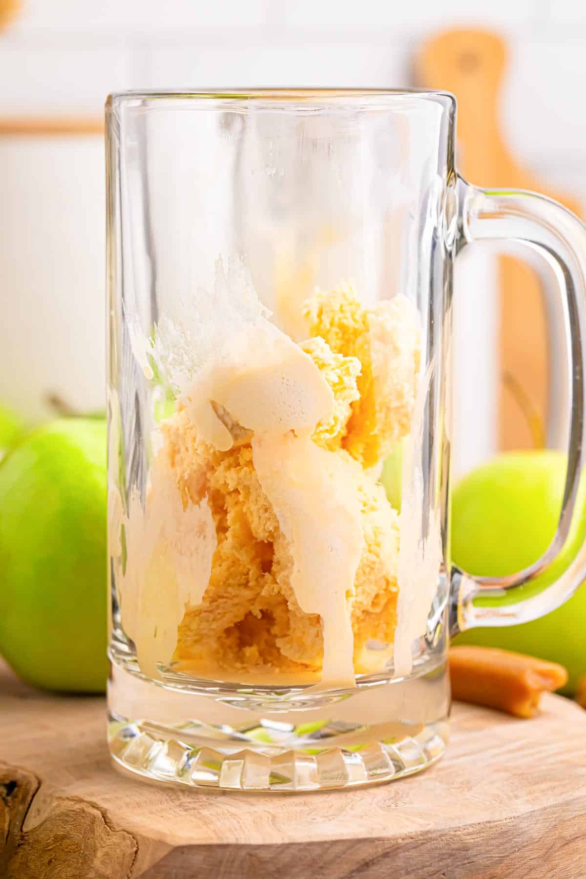 A glass mug filled with caramel and vanilla ice cream on a wooden table with green apples in the background on the counter.