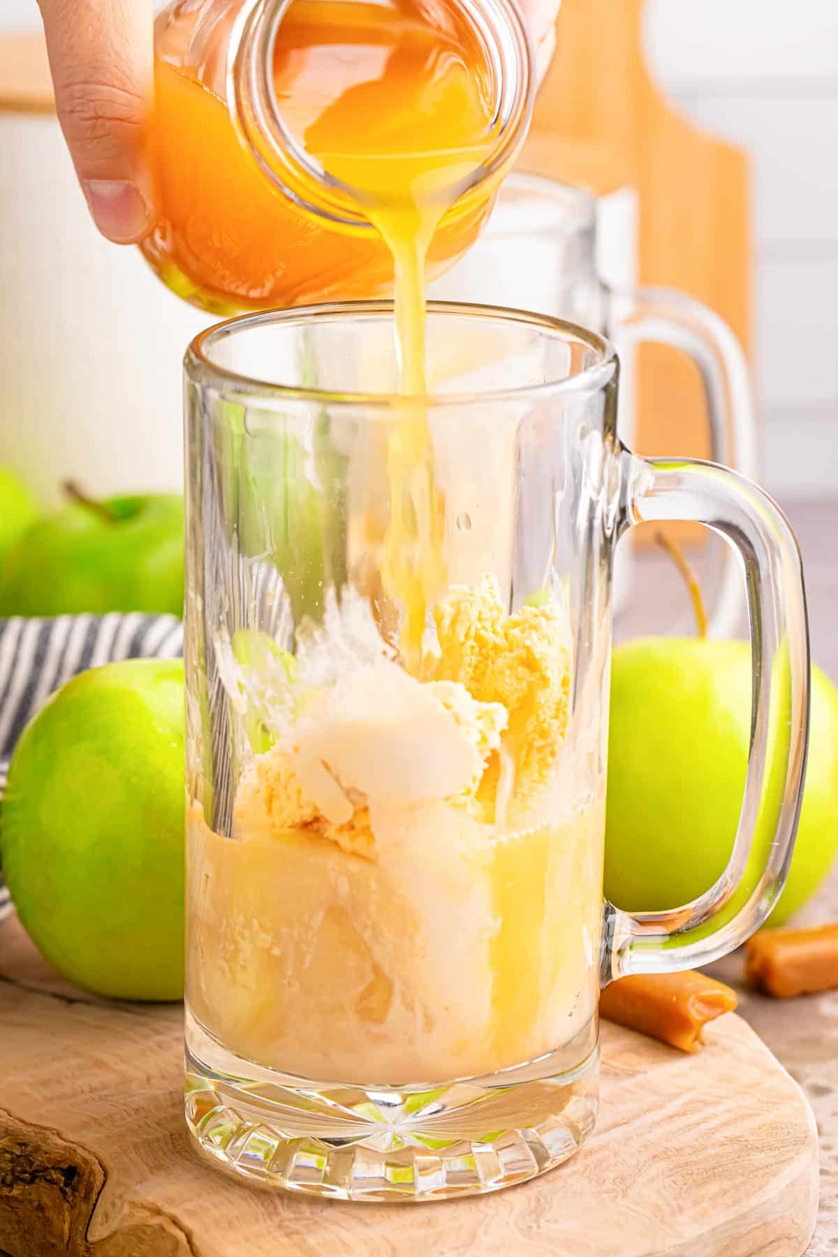 A hand pours apple cider into a glass mug filled with vanilla ice cream with apples in the background on the counter.