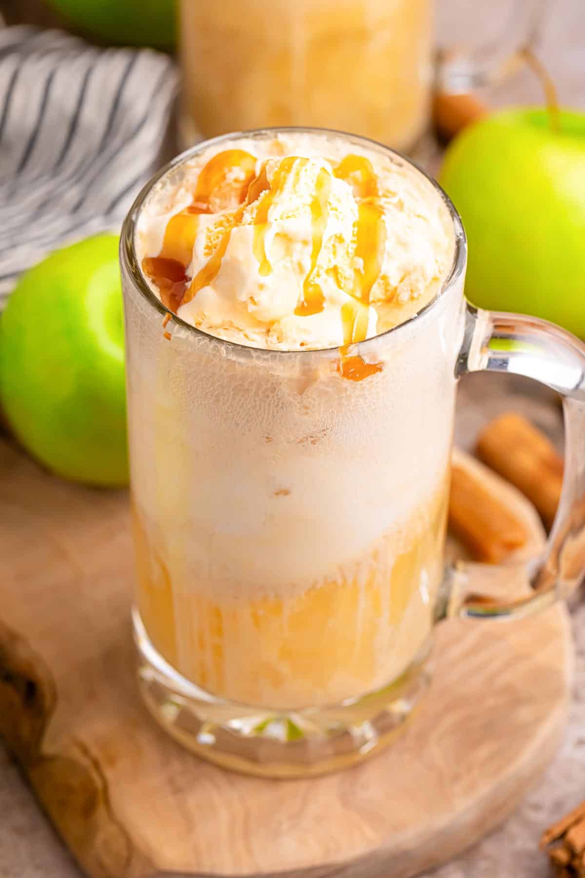 A glass mug filled with an apple cider float topped with whipped cream and caramel drizzle on a piece of wood with apples in the background.