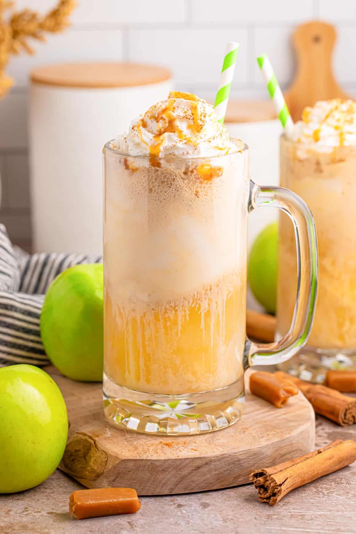 A caramel apple cider float topped with whipped cream and caramel drizzle in a glass mug, with green apples and cinnamon sticks on the counter in the background.