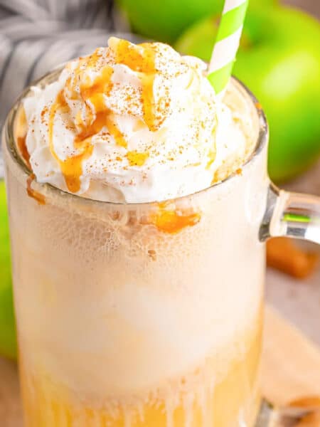 A mug filled with a creamy beverage topped with whipped cream and caramel drizzle, accompanied by a straw. Green apples and a striped cloth are in the background.