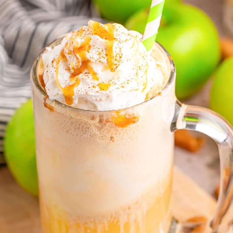 A mug filled with a creamy beverage topped with whipped cream and caramel drizzle, accompanied by a straw. Green apples and a striped cloth are in the background.