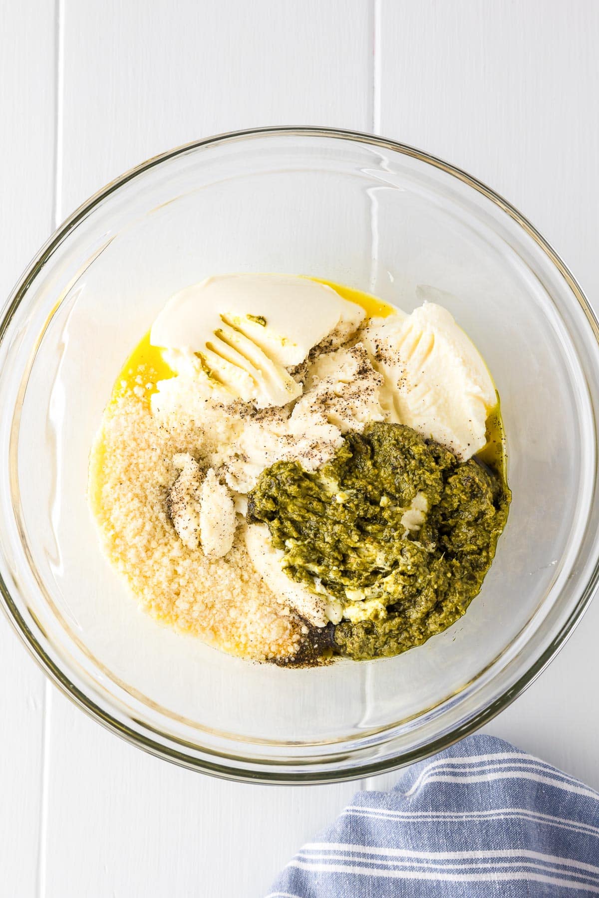 A clear glass bowl full of unmixed ingredients including parmesan cheese, ricotta cheese, pesto sauce, and black pepper for baked rigatoni.