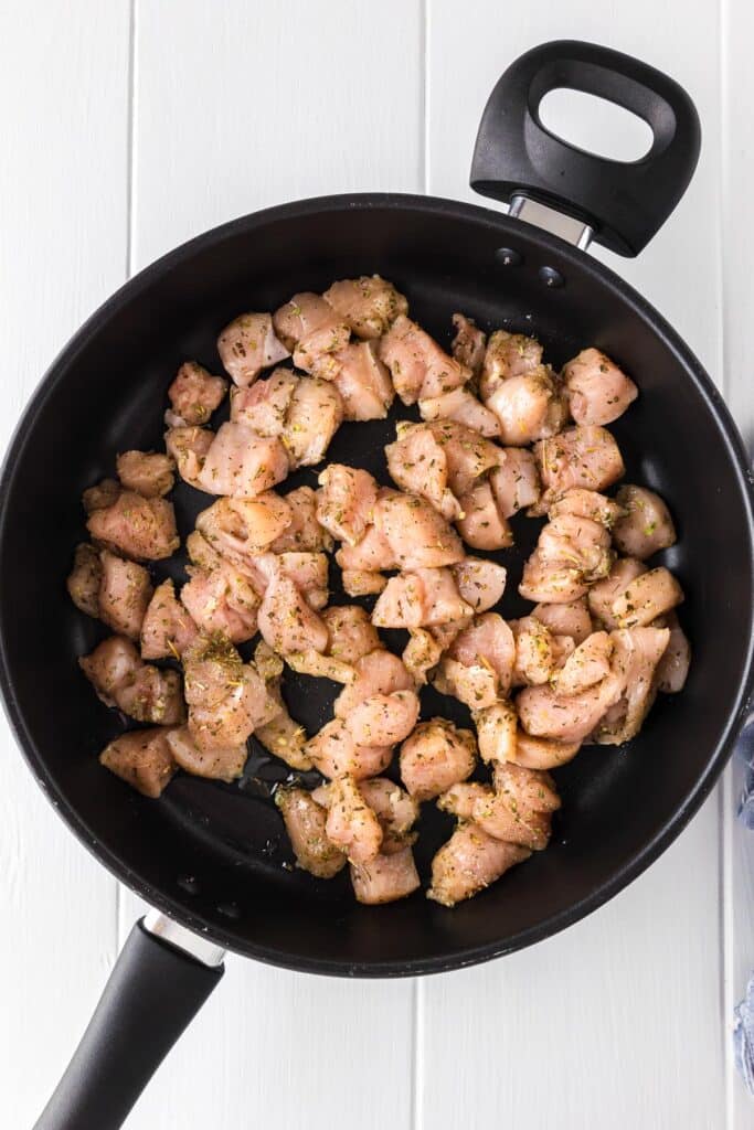 Diced, seasoned raw chicken being cooked in a large skillet from above.