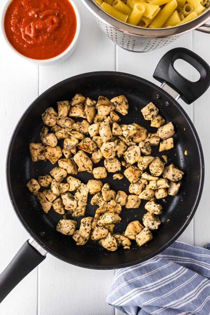 A large black skillet with cooked, seasoned chicken pieces with a bowl of marinara and a large pot full of cooked rigatoni pasta nearby.