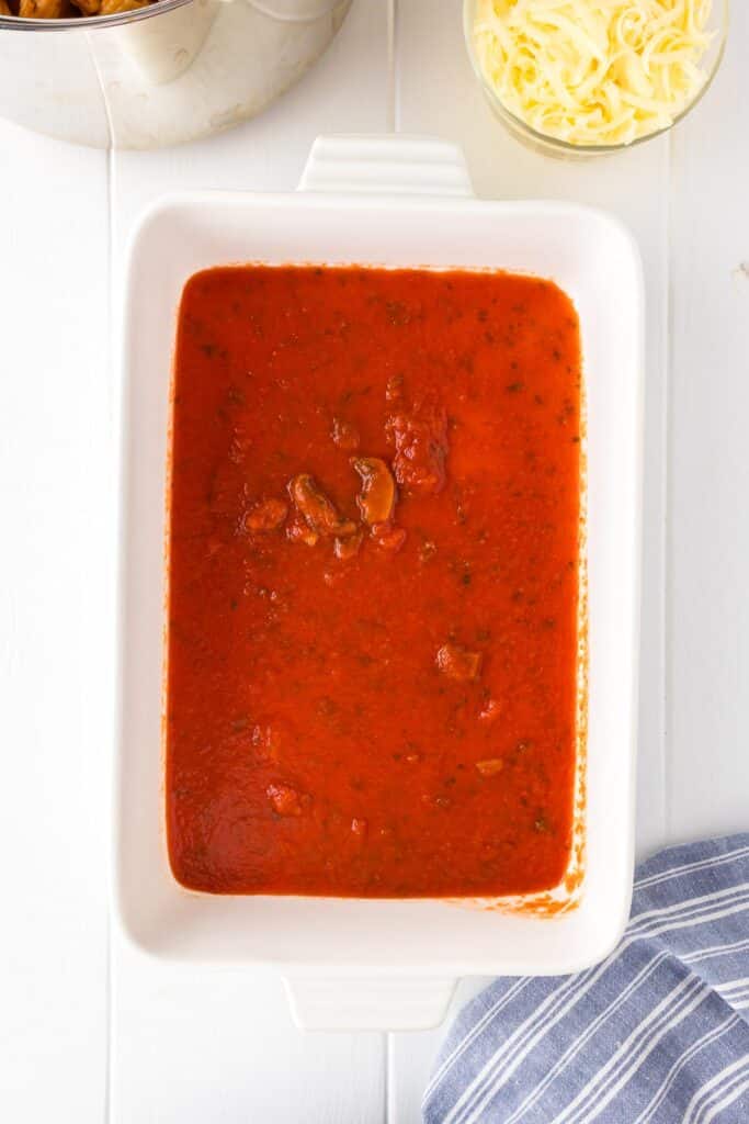 A rectangular baking dish is filled with red tomato sauce on the bottom with a pot full of rigatoni pasta and chicken covered in sauce and shredded mozzarella cheese nearby.