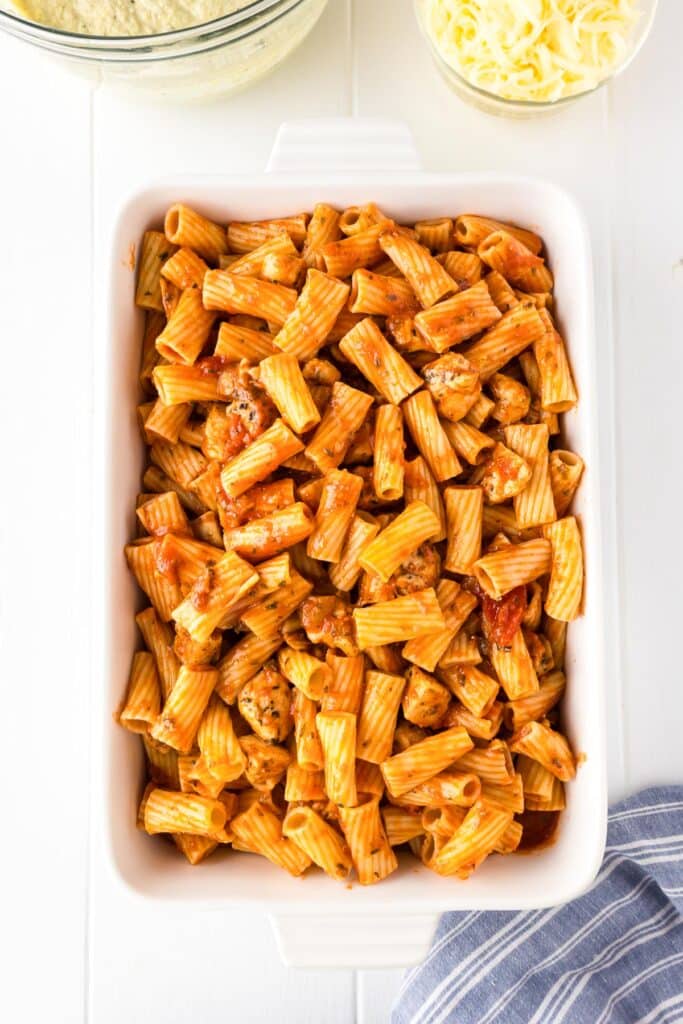 A white rectangular dish filled with rigatoni pasta and chicken coated in a red marinara sauce.