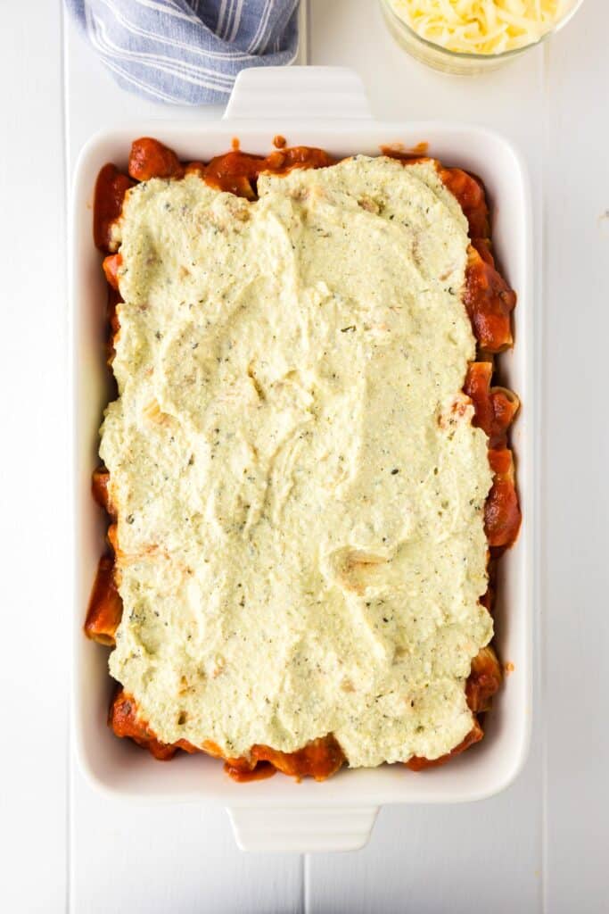 A white rectangular baking dish filled with layers of rigatoni pasta, chicken, tomato sauce, and a thick layer of ricotta cheese mixture spread on top.