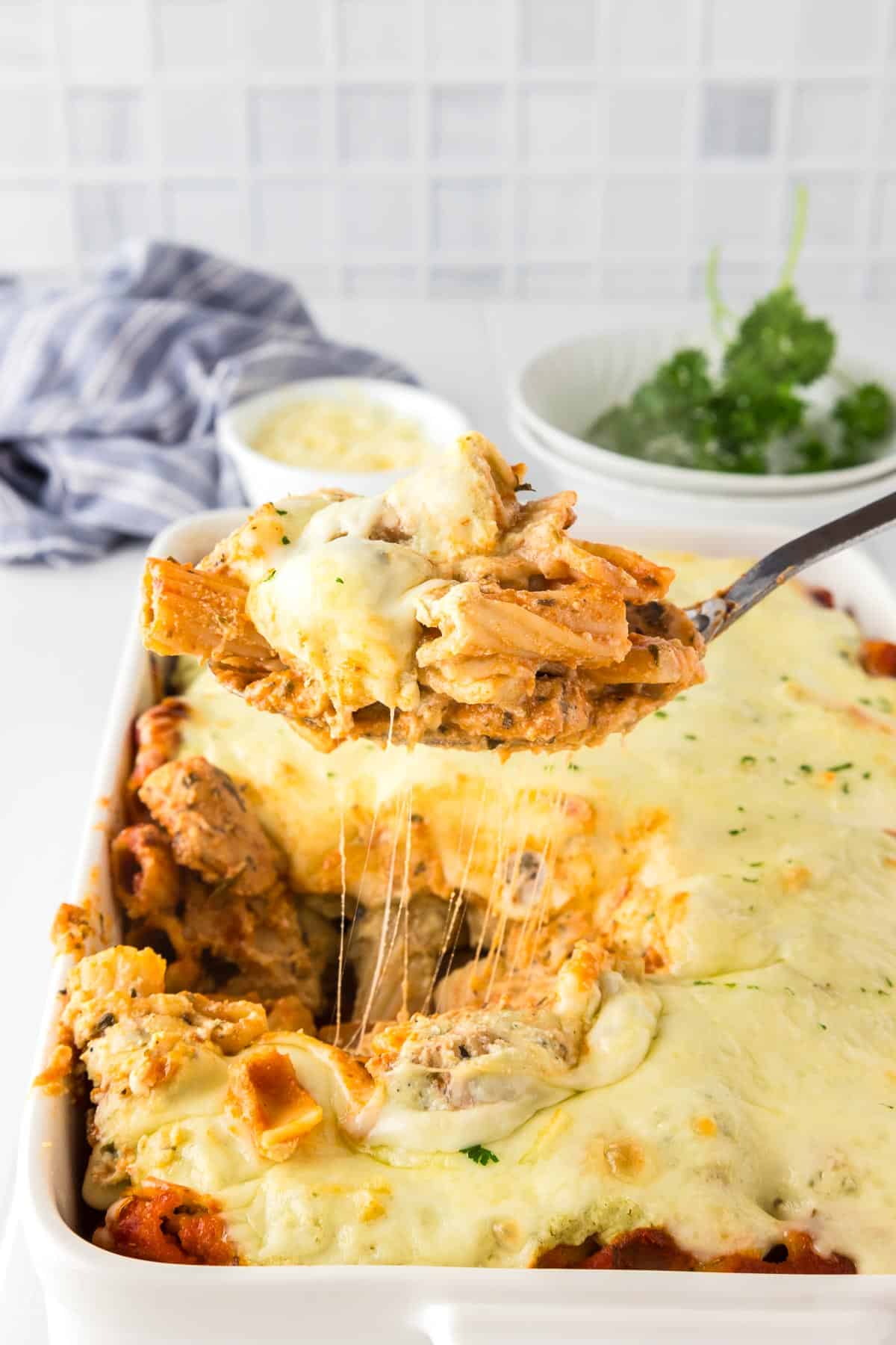 A serving spoon lifting a portion of cheesy baked rigatoni from a casserole dish, with melted cheese stretching.