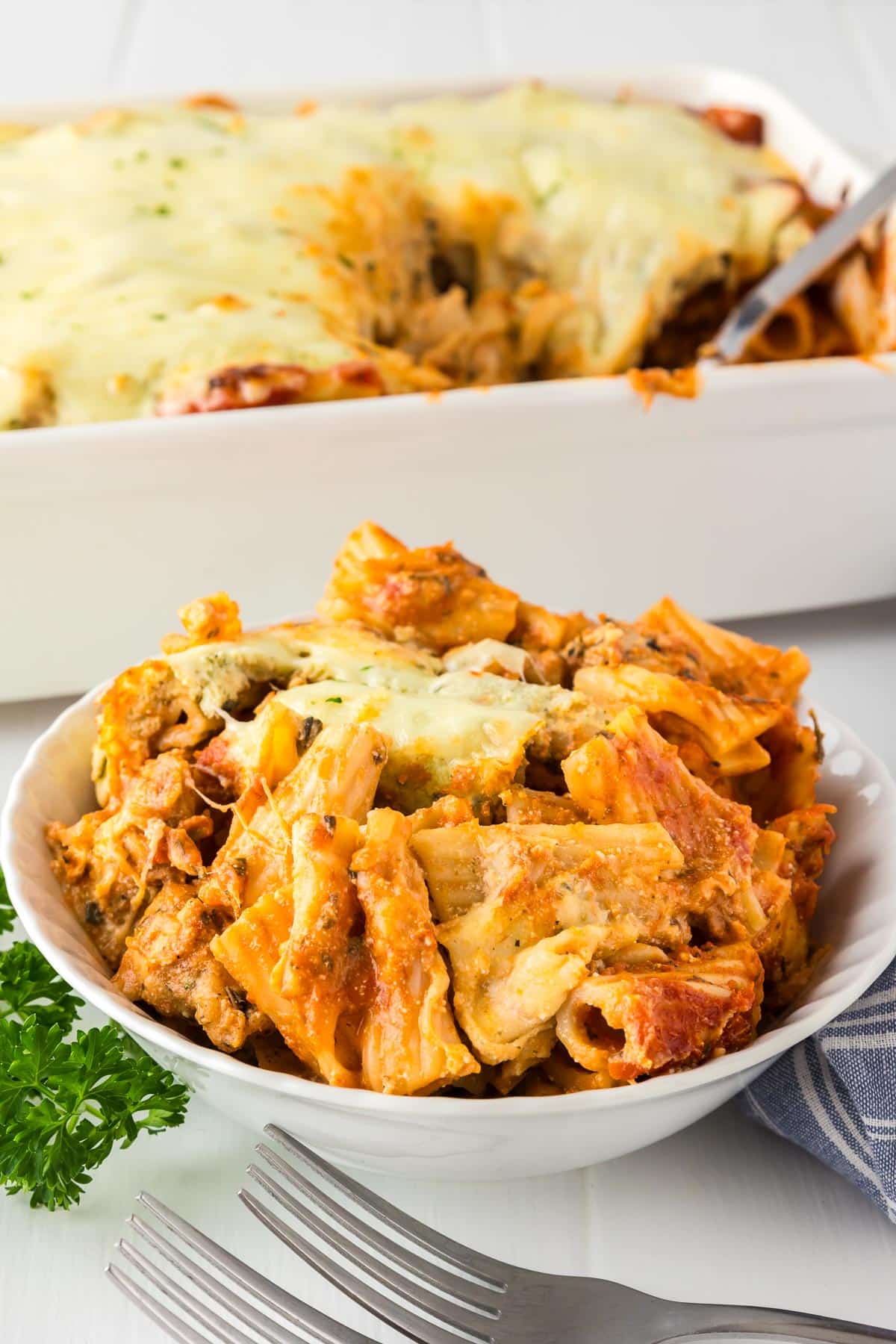 A bowl of baked rigatoni with chicken and cheese next to a baking dish in the background filled with more pasta bake.