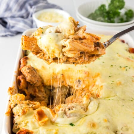 A serving spoon lifts a portion of cheesy baked rigatoni with chicken from a baking dish with the mozzarella cheese pulling.