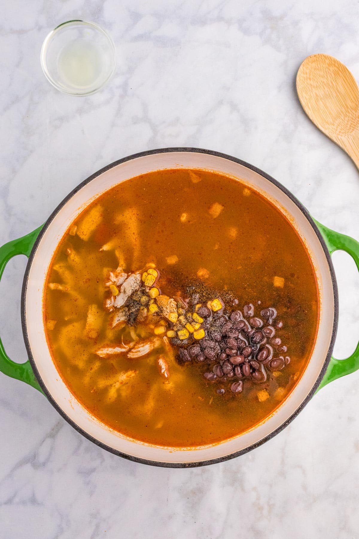 A large pot filled with chicken tortilla soup including corn, black beans, and rotisserie chicken pieces in a tomato broth on a marble countertop next to a wooden spoon and a small glass bowl.