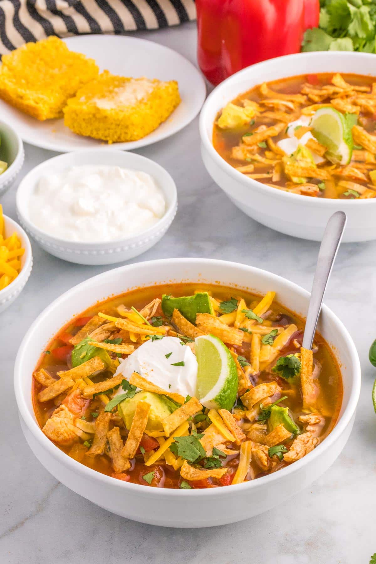 A bowl of chicken tortilla soup garnished with lime, sour cream, cilantro, and shredded cheese with another bowl of soup, a piece of cornbread and more garnish toppings in the background.