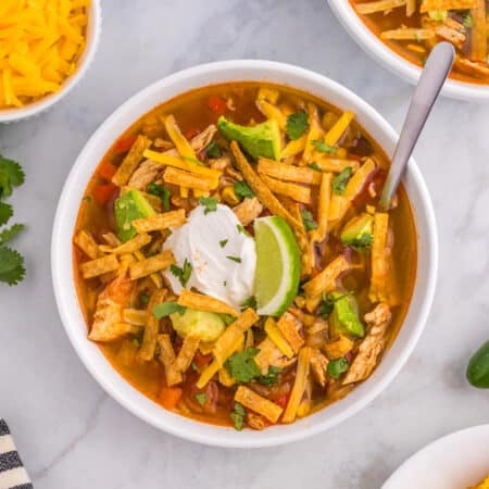 A square view of a bowl of chicken tortilla soup topped with tortilla strips, sour cream and a slice of lime.