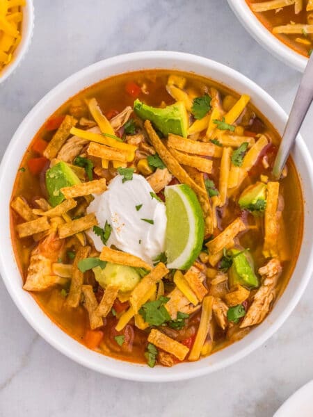 A square view of a bowl of chicken tortilla soup topped with tortilla strips, sour cream and a slice of lime.