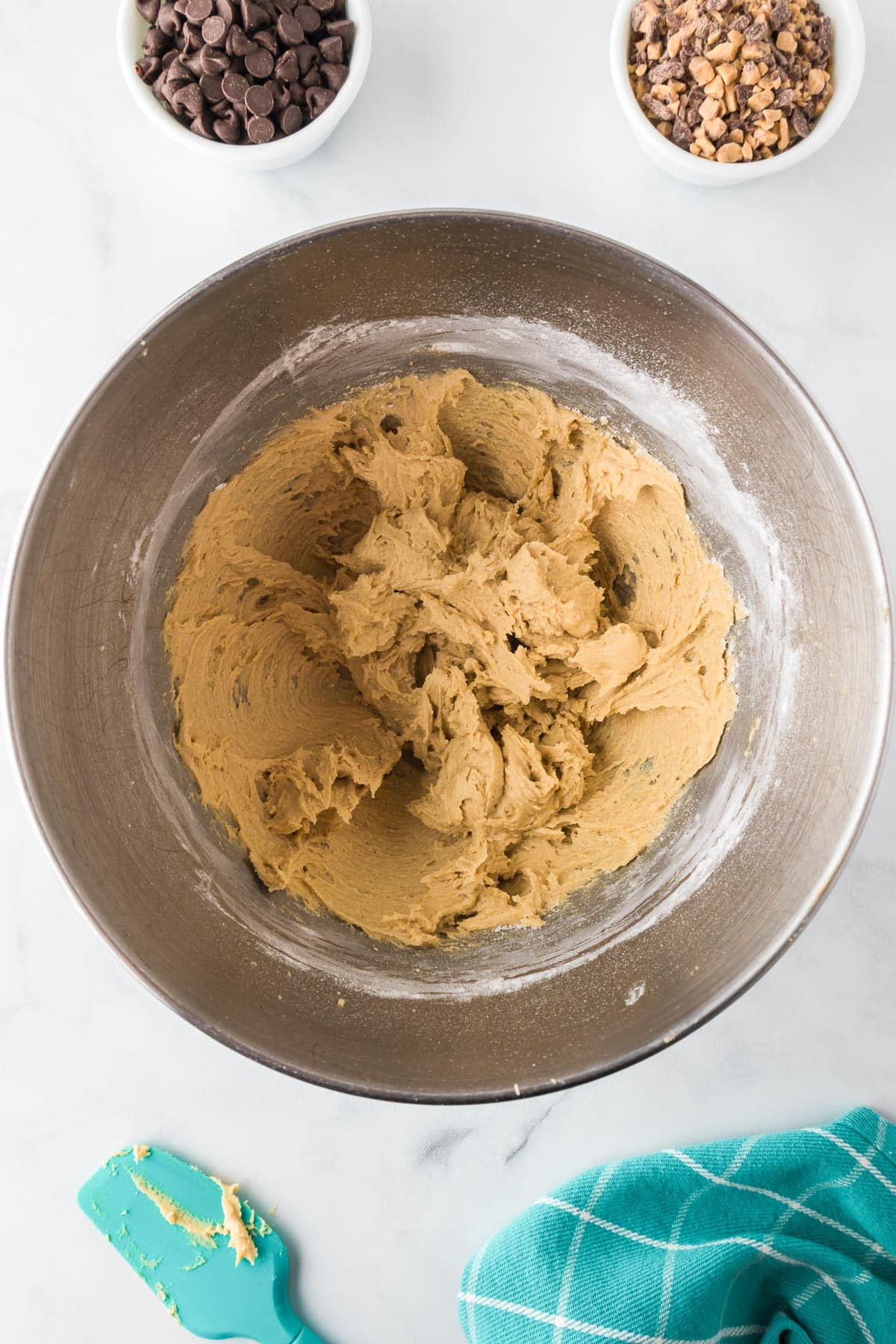 A mixing bowl containing full of cookie dough mixture, with small bowls of chocolate chips and toffee bits in bowls nearby on the counter.