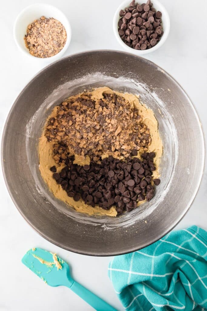 A mixing bowl containing cookie dough, chocolate chips, and chopped toffee bits being mixed in with a spatula.