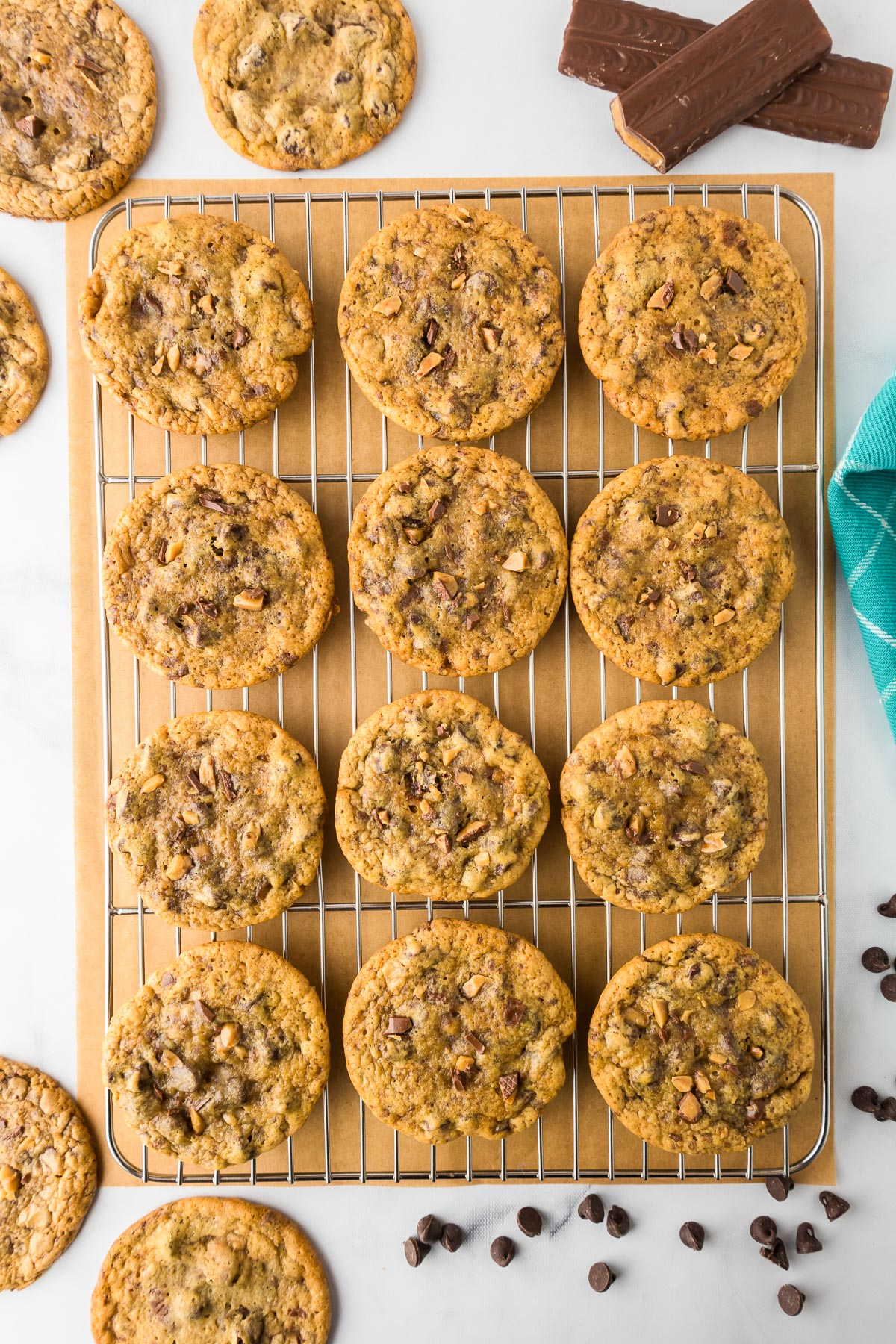 A cooling rack with a dozen chocolate chip toffee cookies lined up with parchment paper underneath and more cookies, chocolate chips and large toffee pieces on the counter nearby.