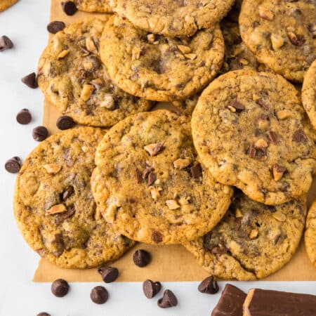 Chocolate chip toffee cookies piled high on parchment paper with chocolate chips scatted on the counter nearby.