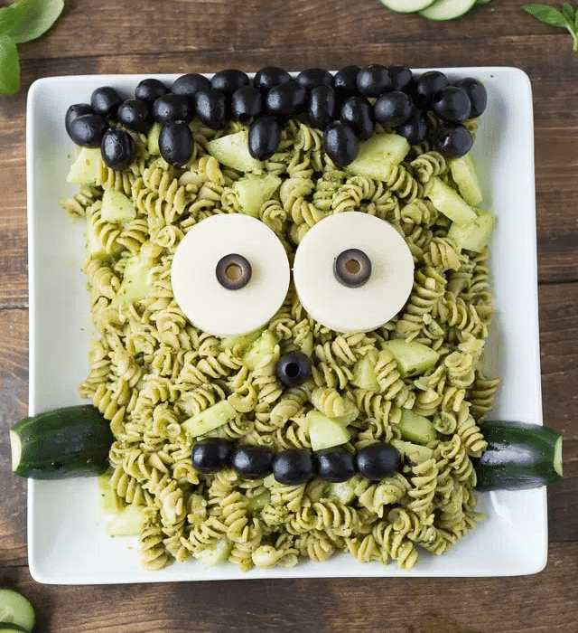 A plate of green pasta salad arranged to resemble a Frankenstein face with olives and cheese on a platter.