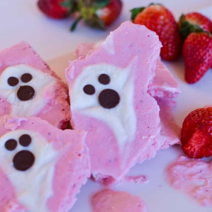 Three pieces of pink yogurt bark with white ghost shapes and chocolate chip eyes and mouths piled next to fresh strawberries.