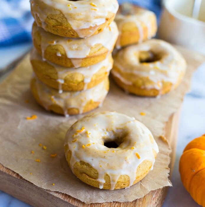 A stack of five glazed pumpkin orange donuts sits on a wooden board, with three additional glazed donuts placed around it.