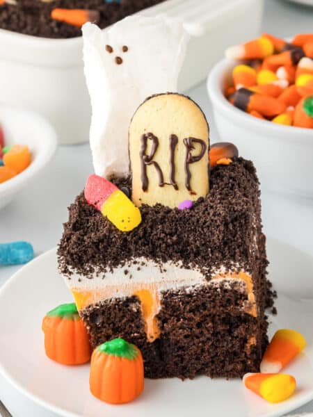 A chocolate Halloween poke cake topped with a a cookie headstone that says RIP, a ghost marshmallow, gummy worms and candy pumpkins.