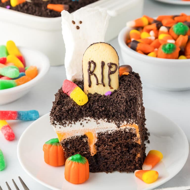 A chocolate Halloween poke cake topped with a a cookie headstone that says RIP, a ghost marshmallow, gummy worms and candy pumpkins.