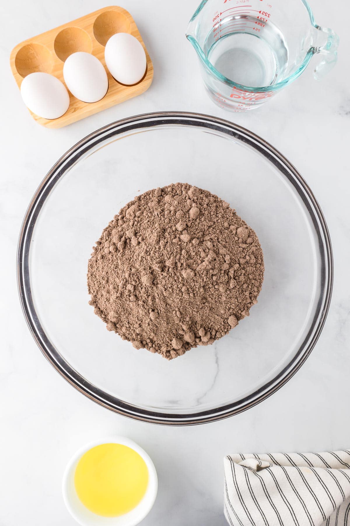 A glass bowl filled with chocolate cake mix with eggs, water and oil in bowls nearby for the Halloween poke cake.