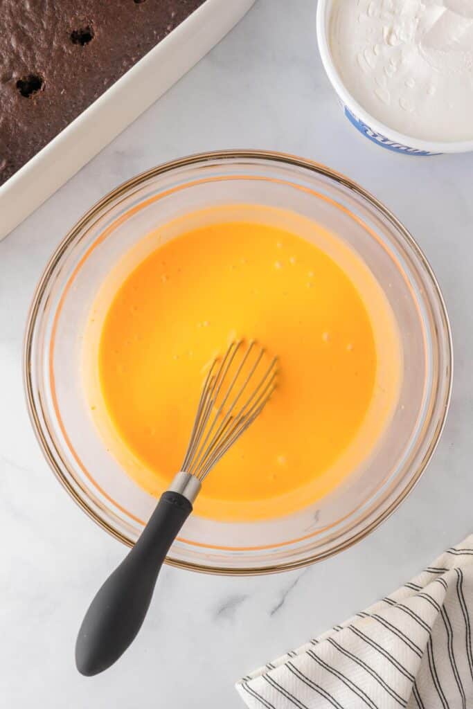 A bowl of pudding being mixed and dyed orange for the Halloween poke cake in a pan nearby.