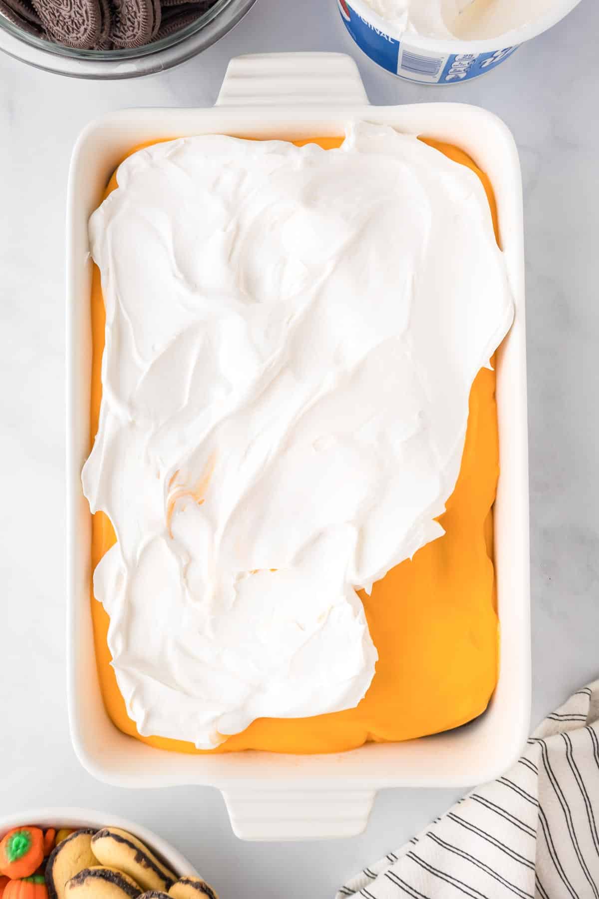 A rectangular baking dish filled with a Halloween poke cake covered in an orange pudding layer with a cool whip frosting layer being added.