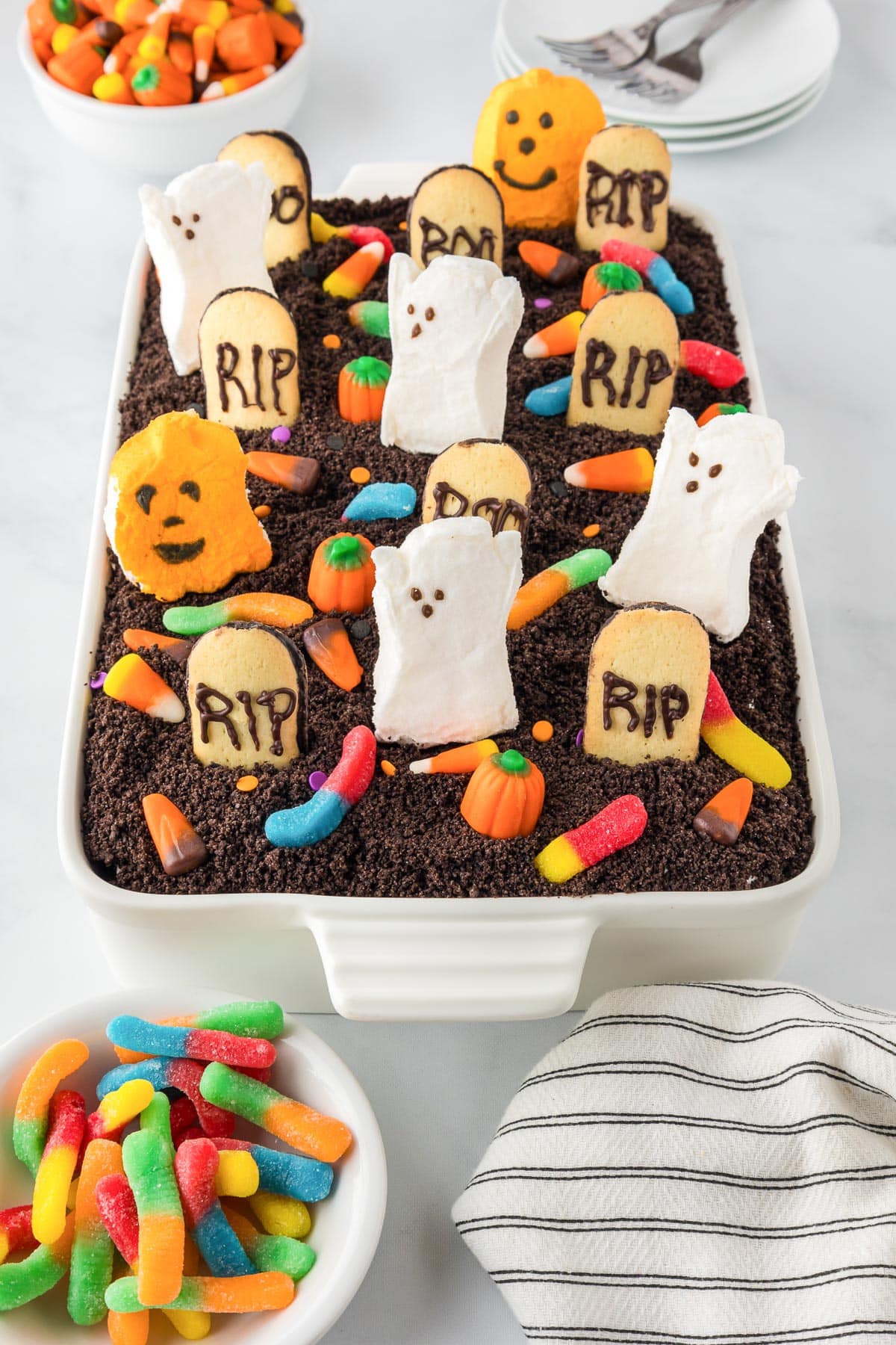 A Halloween poke cake decorated to look lik a graveyard with cookie headstones and marshmallow ghosts and pumpkins from the edge.