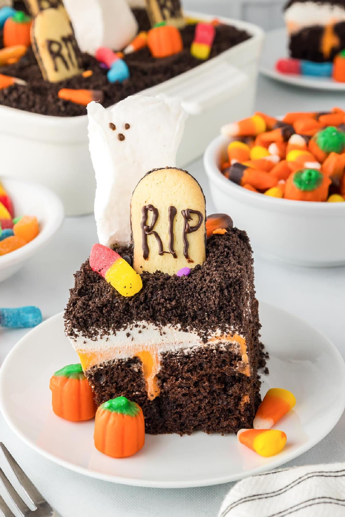 A chocolate Halloween poke cake slice on a plate topped with a a cookie headstone that says RIP, a ghost marshmallow, gummy worms and candy pumpkins with the full pan of cake and more candy in the background.