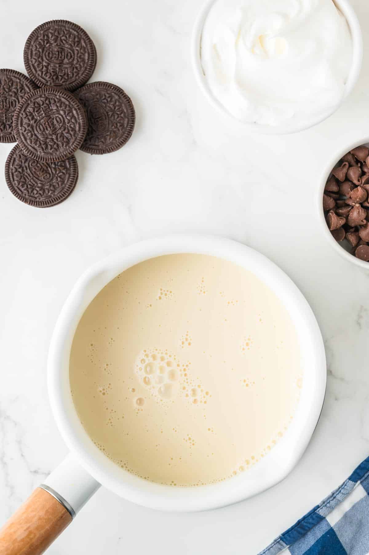 A pot filled with milk surrounded by Oreo cookies, whipped cream and chocolate chips for Oreo hot chocolate.