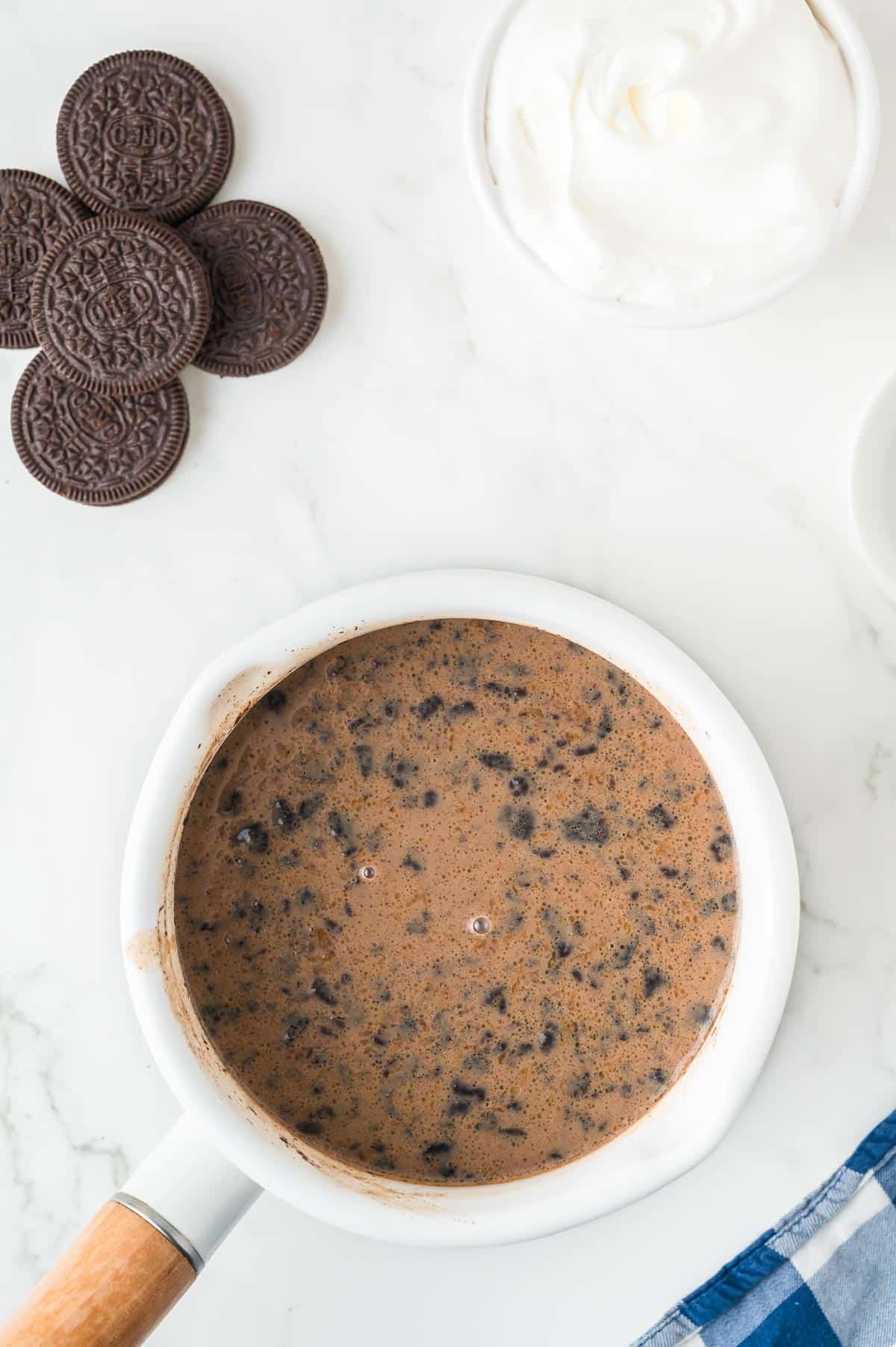 A pot full of a mixture of milk, melted chocolate chips and Oreo cookie crumbs with more Oreo cookies and whipped cream on the counter nearby.