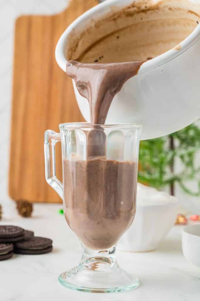 Oreo hot chocolate being poured from a large pot into a large glass handled glass.