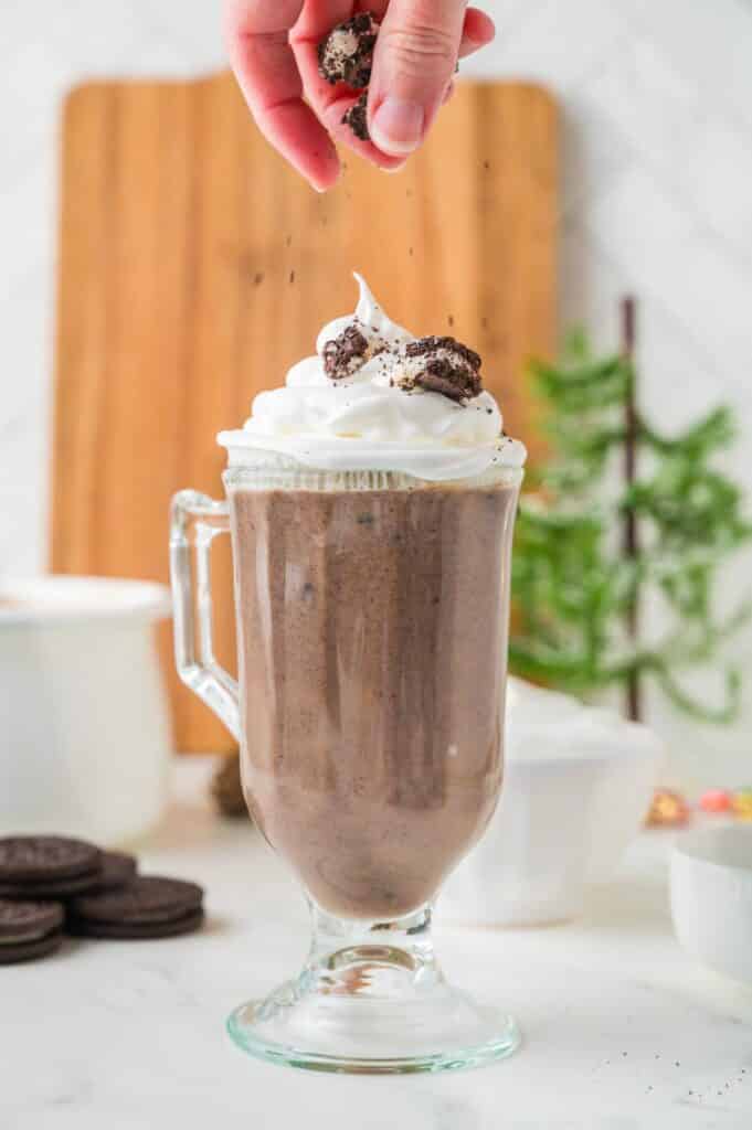 A hand sprinkles crushed Oreo cookies onto whipped cream on top of a glass of Oreo hot chocolate.