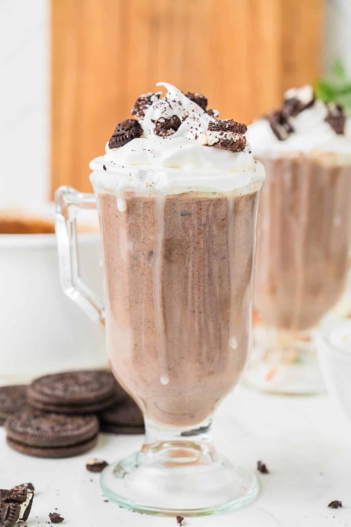 A glass of Oreo hot chocolate topped with whipped cream and crushed Oreo cookies with more cookies and a second glass of hot chocolate in the background.