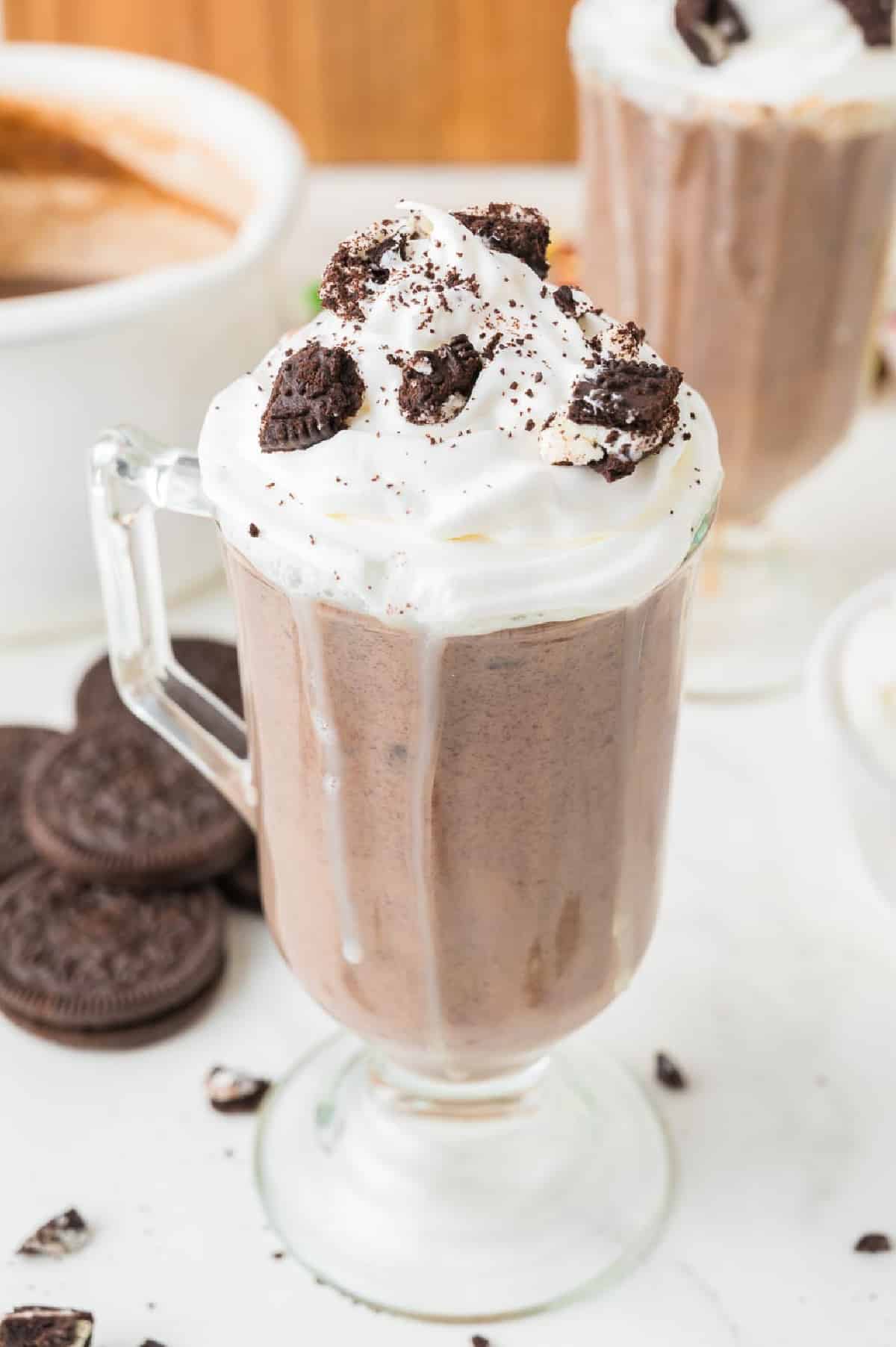 A glass mug filled with Oreo hot chocolate from overhead with dribbles down the glass and more Oreo cookies, the pot from the hot chocolate and a second full glass of Oreo hot chocolate in the background.