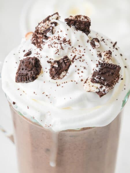 A square view of a glass mug filled with Oreo hot chocolate topped with whipped cream and crushed cookies.