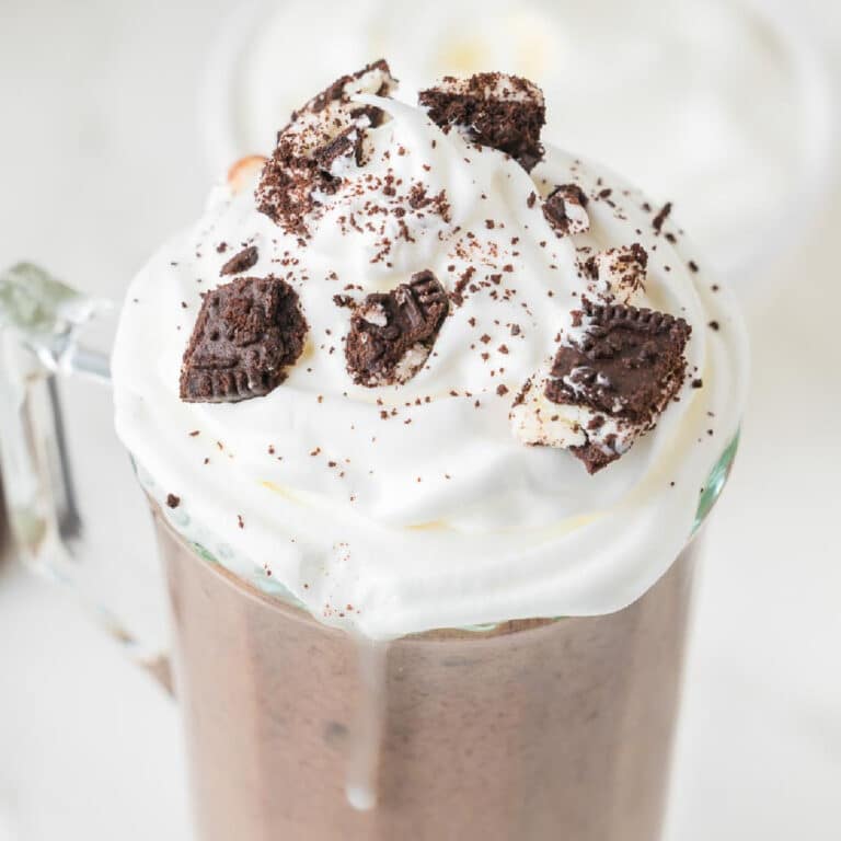 A square view of a glass mug filled with Oreo hot chocolate topped with whipped cream and crushed cookies.