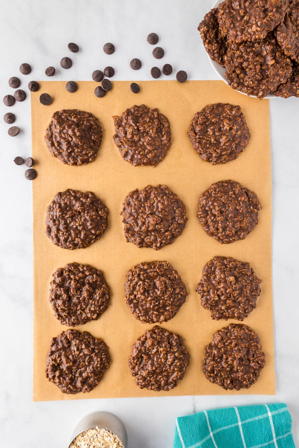 A dozen no bake chocolate peanut butter cookies on parchment paper cooling with more cookies nearby in a bowl.