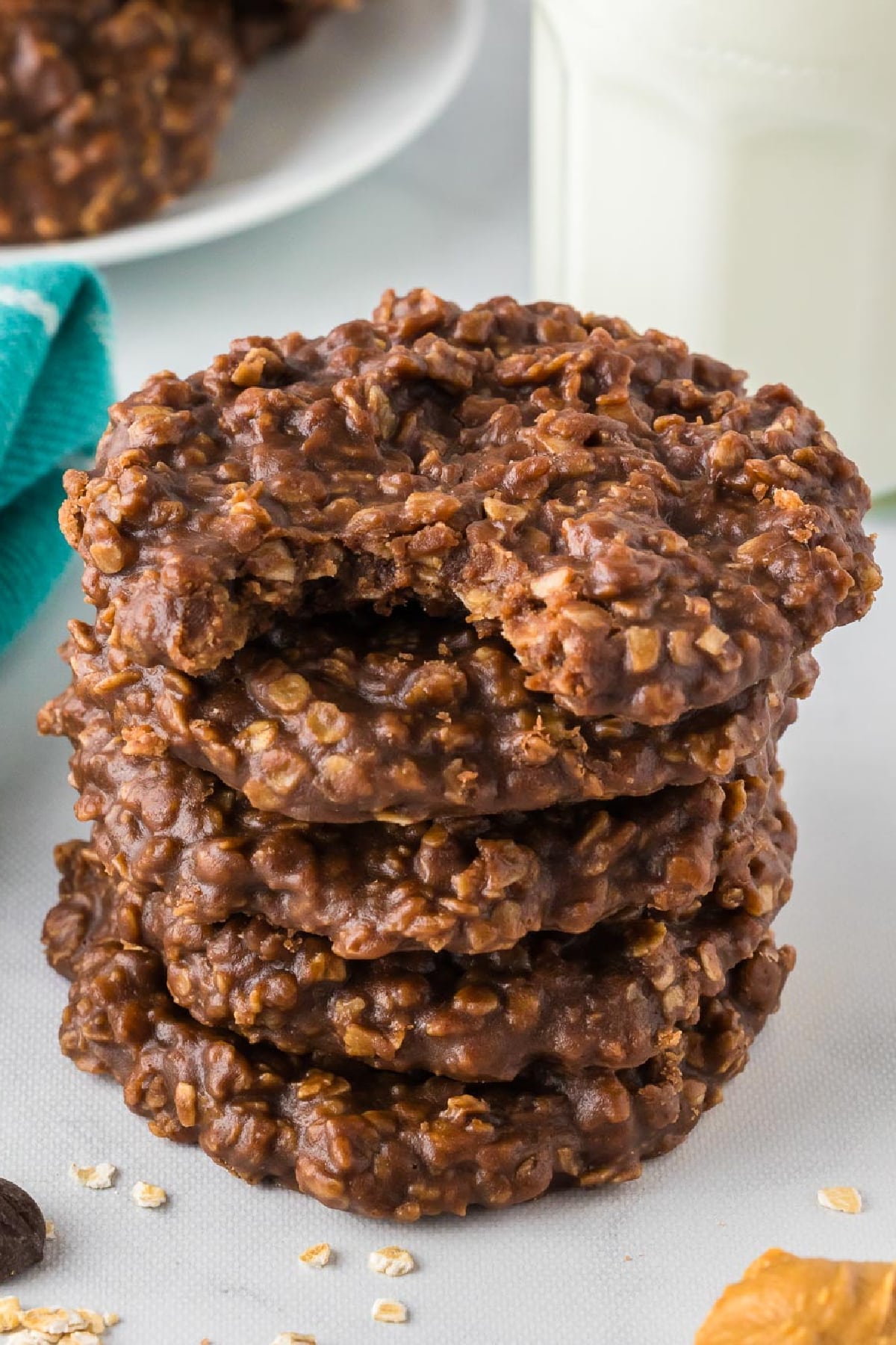 A stack of six chocolate oatmeal cookies, with a bite taken out of the top cookie. A teal cloth and a glass of milk are in the background.